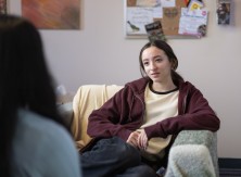 A young person with long hair sits in an armchair looking towards the camera.