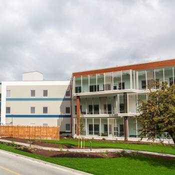 The Red Fish Healing Centre: A modern three-storey building with big glass windows, surrounded by grass and trees, with a two-lane road running in front.