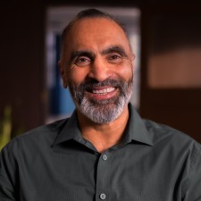 Aman, a South Asian man, smiles at us. He has dark hair and a grey beard, and he wears a dark grey button-up. 