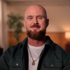 Chris, a man in his 30s or 40s, smiles at us. He is bald, with light skin and a brown beard, and wears an open work shirt over a white t-shirt with a necklace.