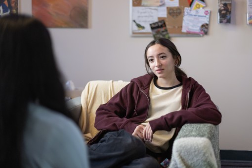 A young person with long hair sits in an armchair looking towards the camera.