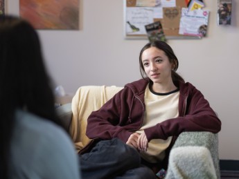 A young person with long hair sits in an armchair looking towards the camera.